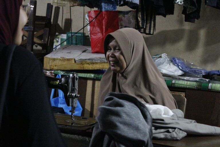 Ibu Titik, seorang penjahit senior berhijab cokelat, sedang fokus menjahit pakaian menggunakan mesin jahit di kiosnya yang berada di lantai dua Pasar Besar Malang.